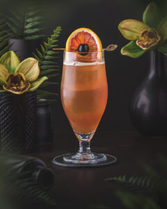 Singapore Sling - orange red classic cocktail served in a pilsner glass on ice, garnished with a lemon wheel and cocktail cherry