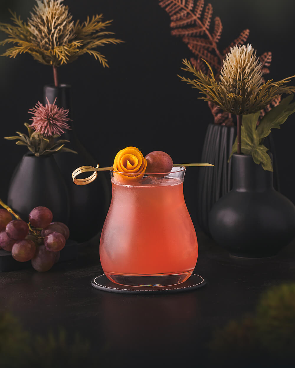 Fall Enzoni - Red orange Cocktail made with bourbon whiskey, Campari and red grapes. Served in a tumbler garnished with an orange lemon peel flower and a red grape.